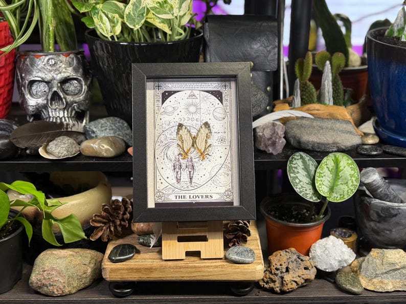 May include: A framed tarot card print of "The Lovers" with a black frame, displayed on a wooden easel. The card features a detailed illustration of two figures with butterfly wings, set against a celestial background. The image is surrounded by various decorative items.