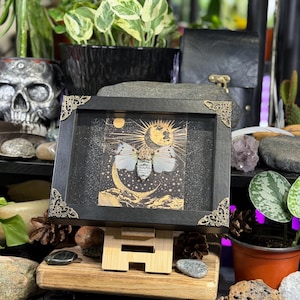 May include: A framed artwork featuring a moth with a celestial design. The black frame has ornate corner details. The artwork depicts a moth with a moon and sun design in gold and white. The artwork is displayed on a wooden easel.