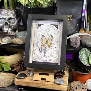 May include: A framed tarot card print featuring a celestial design with the words "THE LOVERS" at the bottom. The artwork includes illustrations of butterflies and figures. The frame is black, and the print is displayed on a small wooden easel.