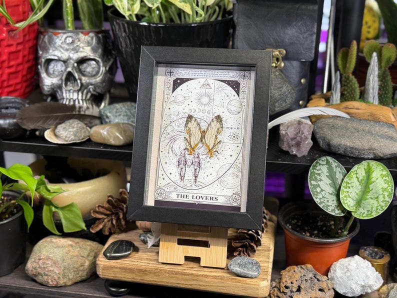 May include: A framed tarot card print featuring a celestial design with the words "THE LOVERS" at the bottom. The artwork includes illustrations of butterflies and figures. The frame is black, and the print is displayed on a small wooden easel.