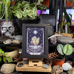 Pu&ograve; includere: Una stampa incorniciata della carta dei tarocchi "The Star", con un design celeste e una figura alata. L'opera d'arte &egrave; in una cornice nera, esposta su un cavalletto di legno, circondata da piante e oggetti decorativi. Il testo "THE STAR" &egrave; in basso.