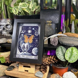 May include: A framed print of "The Lovers" tarot card, featuring a celestial design with a butterfly and two figures. The artwork is in a black frame, displayed on a wooden easel, surrounded by plants and decorative items. The print has a blue background with white illustrations.
