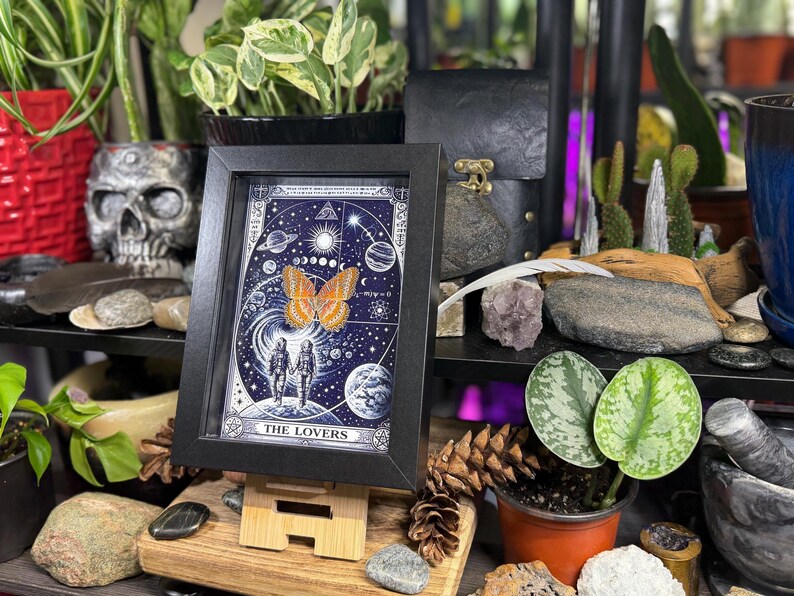 May include: A framed print of "The Lovers" tarot card, featuring a celestial design with a butterfly and two figures. The artwork is in a black frame, displayed on a wooden easel, surrounded by plants and decorative items. The print has a blue background with white illustrations.