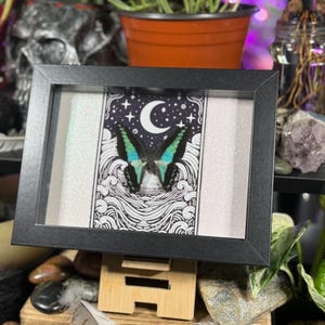 May include: A framed art piece featuring a butterfly with teal and black wings, set against a night sky design with a crescent moon and stars. The artwork is in a black frame, displayed on a wooden stand.