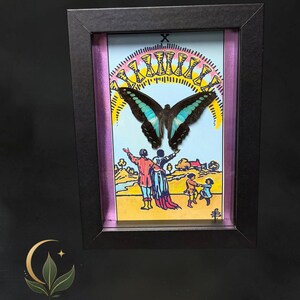 Puede incluir: Un marco negro tipo caja que contiene una ilustraci&oacute;n de la carta del tarot Diez de Copas, con una mariposa grande de alas negras y turquesas centrada sobre una escena de figuras bajo un arco&iacute;ris de diez copas doradas sobre un cielo azul.