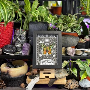 May include: A framed tarot card print titled "The Universe" is displayed on a wooden easel. The black and white artwork features celestial imagery and is surrounded by plants, crystals, and decorative items. A skull and other decorative items are also visible.