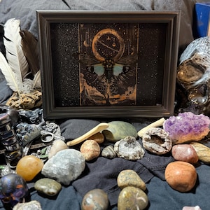 May include: A framed artwork with a celestial moth design and blue wings, set against a dark background. The image is surrounded by crystals, stones, feathers, and a decorative skull, creating a mystical and artistic display.