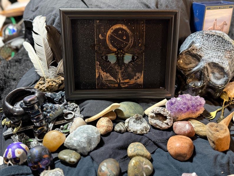 May include: A framed artwork with a celestial moth design and blue wings, set against a dark background. The image is surrounded by crystals, stones, feathers, and a decorative skull, creating a mystical and artistic display.