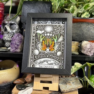 May include: A framed artwork featuring a colorful butterfly against a black and white celestial background. The artwork is labeled "The Universe" and is displayed on a wooden stand. The butterfly has orange, green, and yellow wings. The frame is black.