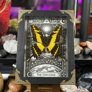 Peut inclure: Une &oelig;uvre encadr&eacute;e repr&eacute;sentant un papillon jaune et noir sur un fond c&eacute;leste en noir et blanc. Le cadre noir est orn&eacute; d'angles dor&eacute;s et le texte "THE UNIVERSE" est visible en bas.
