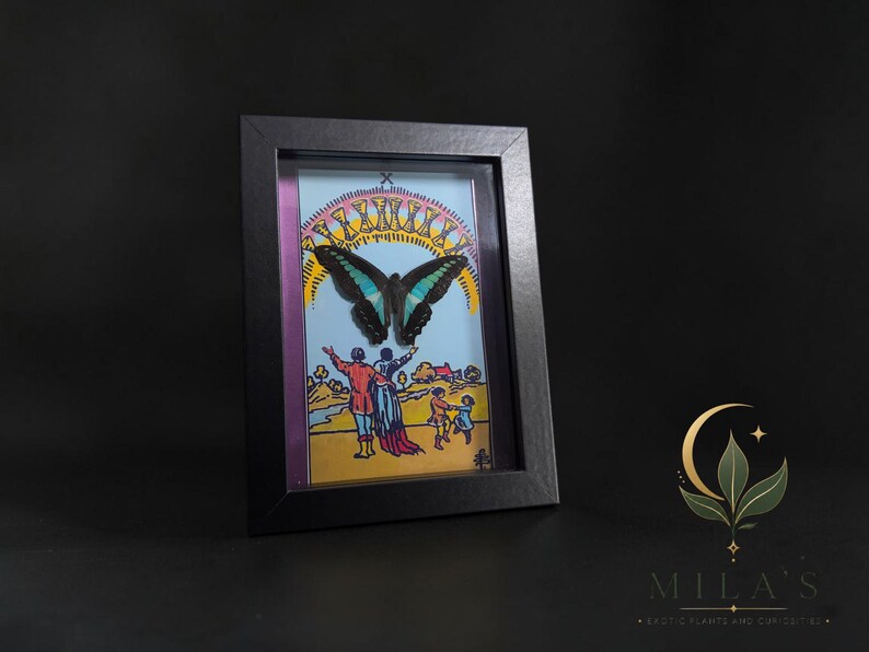 Puede incluir: Una obra de arte enmarcada que presenta una mariposa negra y azul cerceta sobre una colorida ilustraci&oacute;n de figuras en un paisaje. La escena incluye un cielo amarillo con un arco&iacute;ris de diez copas doradas, todo dentro de un marco negro simple.