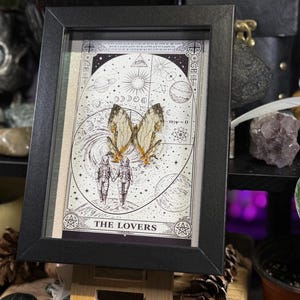 May include: Framed artwork featuring a tarot card design with the title "THE LOVERS." The artwork depicts two figures in space suits, butterfly wings, and celestial elements. The frame is black, and the artwork is displayed on a wooden easel.