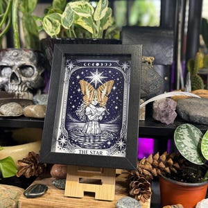 May include: A framed print of "The Star" tarot card, depicting a woman with butterfly wings in water under a starry sky. The artwork is in black, white, and blue, displayed on a wooden easel. The background includes plants and decorative items, creating a mystical atmosphere.