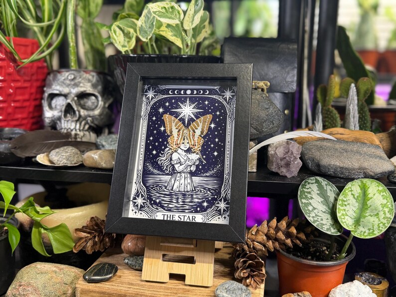 May include: A framed print of "The Star" tarot card, depicting a woman with butterfly wings in water under a starry sky. The artwork is in black, white, and blue, displayed on a wooden easel. The background includes plants and decorative items, creating a mystical atmosphere.
