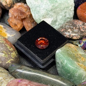 Puede incluir: Una gema facetada, redonda, de color naranja rojizo se exhibe sobre una superficie de terciopelo negro, rodeada de una variedad de rocas y cristales pulidos y coloridos. Las gemas y cristales son en tonos verdes, marrones, naranjas y grises.