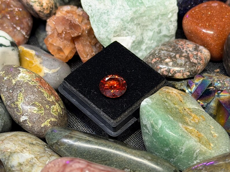 Puede incluir: Una gema facetada, redonda, de color naranja rojizo se exhibe sobre una superficie de terciopelo negro, rodeada de una variedad de rocas y cristales pulidos y coloridos. Las gemas y cristales son en tonos verdes, marrones, naranjas y grises.