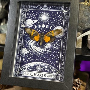 May include: Framed artwork featuring a moth with orange and black wings, set against a dark blue celestial background with planets, stars, and the word "CHAOS". The artwork is in a black frame.