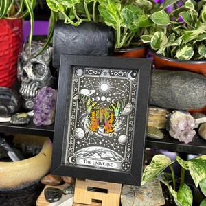 May include: A framed print with a black border, featuring a celestial design with a butterfly and the words "The Universe." The artwork is predominantly black and white with accents of yellow, orange, and green. The print is displayed on a wooden easel.