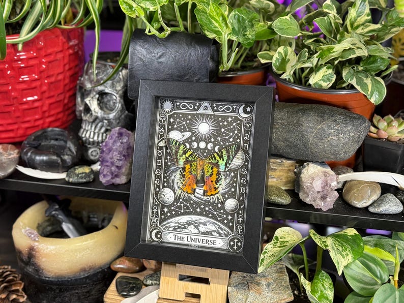 May include: A framed print with a black border, featuring a celestial design with a butterfly and the words "The Universe." The artwork is predominantly black and white with accents of yellow, orange, and green. The print is displayed on a wooden easel.