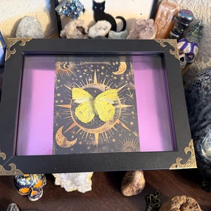 May include: A framed artwork featuring a yellow butterfly against a celestial background with moons and stars. The black frame has decorative gold-colored corner accents, and the background is a vibrant purple. The artwork is displayed among crystals and other decorative objects.
