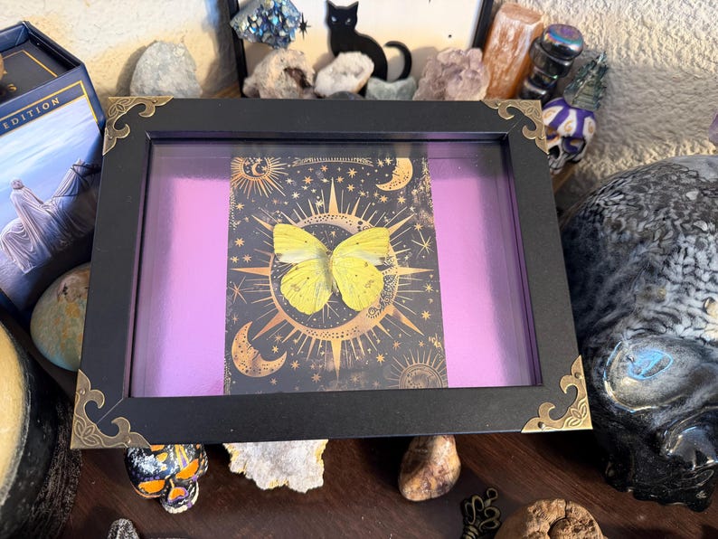 May include: A framed artwork featuring a yellow butterfly against a celestial background with moons and stars. The black frame has decorative gold-colored corner accents, and the background is a vibrant purple. The artwork is displayed among crystals and other decorative objects.