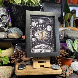 May include: A framed black and white print depicting planets, moon phases, and a butterfly design. The word "CHAOS" is at the bottom. The print is displayed on a small wooden easel, surrounded by plants and decorative items.