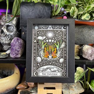 May include: A framed black and white print of "The Universe" tarot card. The artwork features a butterfly with colorful wings, celestial bodies, and the card's title. The frame is displayed on a wooden stand, surrounded by plants and crystals.