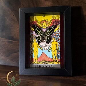 May include: A black rectangular shadow box frame containing a tarot card titled THE LOVERS with the Roman numeral VI. A large, dark-coloured butterfly with light green spots is mounted over the central figure of the card, which depicts two nude figures standing before a mountain and a large yellow sun.