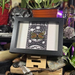 May include: A framed print with a celestial theme, showcasing a butterfly in the centre. The artwork is mainly black and white, with green, orange, and yellow highlights. The frame is black, and the print is displayed on a wooden stand.