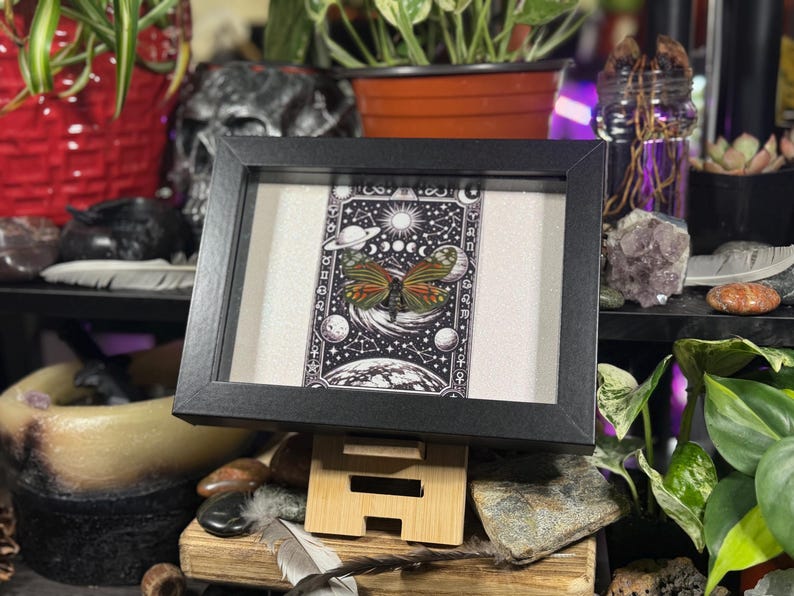 May include: A framed print with a celestial theme, showcasing a butterfly in the centre. The artwork is mainly black and white, with green, orange, and yellow highlights. The frame is black, and the print is displayed on a wooden stand.