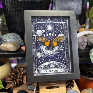 May include: Framed art print featuring a celestial design with a butterfly and planets against a dark blue background. The artwork includes the word "CHAOS" at the bottom and is encased in a black frame. The print is displayed on a wooden easel.