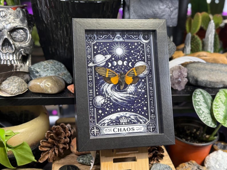 May include: Framed art print featuring a celestial design with a butterfly and planets against a dark blue background. The artwork includes the word "CHAOS" at the bottom and is encased in a black frame. The print is displayed on a wooden easel.