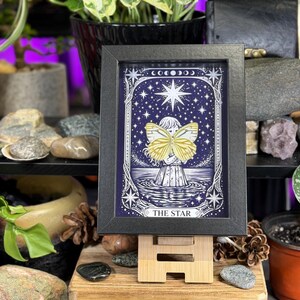 May include: A framed artwork with a celestial theme, depicting a figure with a butterfly against a dark blue starry backdrop. The text "THE STAR" is visible. The black framed print is displayed on a small wooden easel, suitable for a desk or shelf.