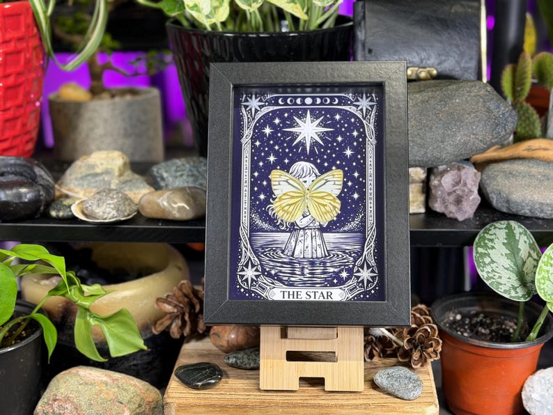 May include: A framed artwork with a celestial theme, depicting a figure with a butterfly against a dark blue starry backdrop. The text "THE STAR" is visible. The black framed print is displayed on a small wooden easel, suitable for a desk or shelf.