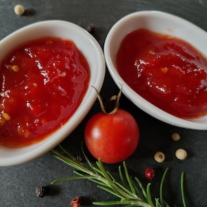 Puede incluir: Dos cuencos ovalados blancos llenos de salsa de tomate roja, un tomate rojo fresco y ramitas de romero. Granos de pimienta y pequeñas semillas blancas dispersas. La imagen está sobre un fondo oscuro.