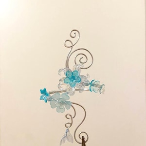 May include: A decorative silver metal floral hair accessory. The piece features clear and turquoise blue flowers, with a delicate chain and a dangling, translucent, fin-shaped charm. The design includes swirls and curls.