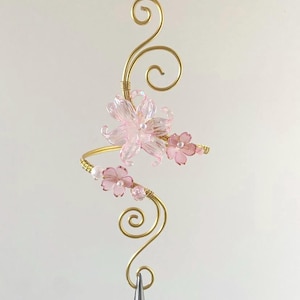 Könnte beinhalten: Ein filigranes, handgefertigtes Haaraccessoire mit floralem Design. Es besteht aus einem goldfarbenen Drahtrahmen mit wirbelnden Akzenten, verziert mit durchscheinenden rosa Blüten und kleinen perlenartigen Perlen. Das Accessoire wird von einer silbernen Pinzette gehalten.