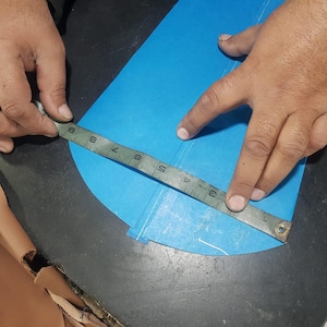 Pu&ograve; includere: Una persona misura un tessuto blu con un righello di metallo. Il righello mostra le misure in centimetri. Il tessuto &egrave; su una superficie scura. Le mani della persona sono visibili.