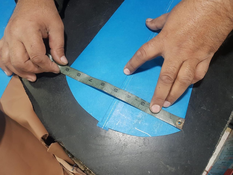 Pu&ograve; includere: Una persona misura un tessuto blu con un righello di metallo. Il righello mostra le misure in centimetri. Il tessuto &egrave; su una superficie scura. Le mani della persona sono visibili.
