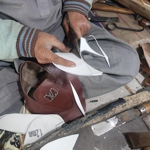 May include: A craftsman cuts a paper pattern for a brown leather shoe. The shoe features a silver buckle. The scene shows the shoemaker at work, surrounded by tools and materials. The shoe has a classic design.