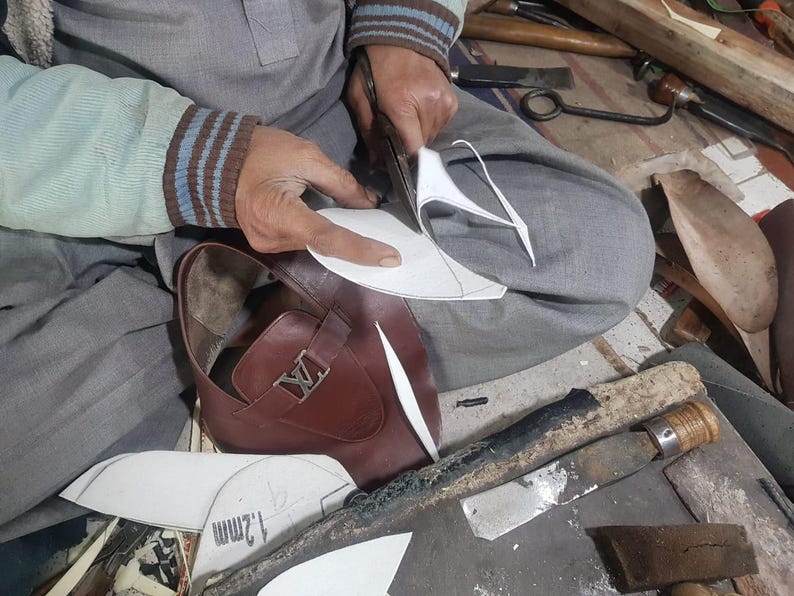 May include: A craftsman cuts a paper pattern for a brown leather shoe. The shoe features a silver buckle. The scene shows the shoemaker at work, surrounded by tools and materials. The shoe has a classic design.
