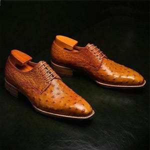 Puede incluir: Un par de zapatos de vestir de cuero marr&oacute;n con una superficie texturizada y un dise&ntilde;o con cordones. Los zapatos tienen un tac&oacute;n bajo y se exhiben con hormas de madera. Los zapatos est&aacute;n sobre una superficie verde oscuro.