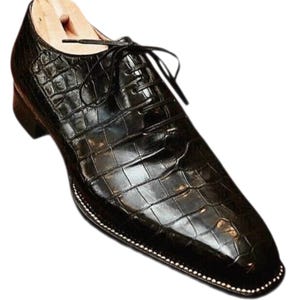 Pu&ograve; includere: Una scarpa da uomo in pelle nera con un motivo strutturato a forma di coccodrillo. La scarpa presenta un design stringato e una punta quadrata. La suola &egrave; delineata da piccole cuciture bianche. All'interno c'&egrave; una forma di scarpa in legno.