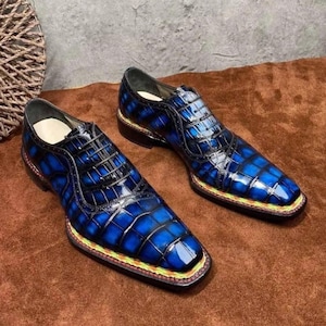 May include: A pair of blue leather dress shoes with a glossy finish. The shoes have a square toe and a textured, crocodile-like pattern. The soles have a colourful trim, and the shoes are laced up with black laces.