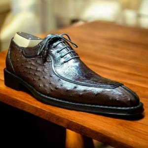 Puede incluir: Zapato de vestir de cuero marrón con un patrón de avestruz texturizado. El zapato presenta un diseño clásico con cordones, una suela negra y una horma de zapato de madera en el interior. El zapato se exhibe sobre una superficie de madera.