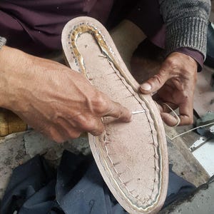 Pu&ograve; includere: Primo piano di una suola di scarpa in fase di realizzazione. La suola &egrave; marrone chiaro con un bordo cucito e piccole borchie metalliche. Le mani di una persona lavorano sulla suola con ago e filo, indicando la fabbricazione o la riparazione di calzature.