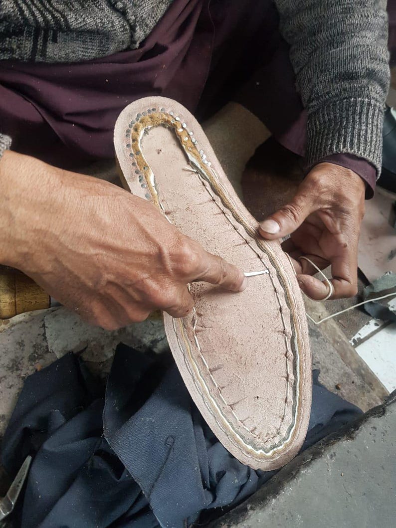 Pu&ograve; includere: Primo piano di una suola di scarpa in fase di realizzazione. La suola &egrave; marrone chiaro con un bordo cucito e piccole borchie metalliche. Le mani di una persona lavorano sulla suola con ago e filo, indicando la fabbricazione o la riparazione di calzature.