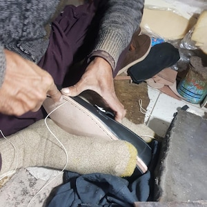 Pu&ograve; includere: Un calzolaio cuce una scarpa in pelle nera. Il calzolaio indossa un maglione grigio e calzini beige. Vari componenti e strumenti per scarpe sono visibili sullo sfondo, suggerendo un'officina. La scarpa &egrave; cucita a mano con filo bianco.