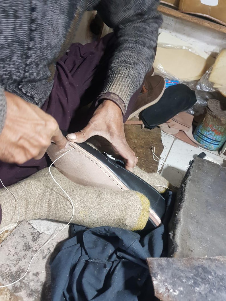 Pu&ograve; includere: Un calzolaio cuce una scarpa in pelle nera. Il calzolaio indossa un maglione grigio e calzini beige. Vari componenti e strumenti per scarpe sono visibili sullo sfondo, suggerendo un'officina. La scarpa &egrave; cucita a mano con filo bianco.
