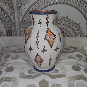 May include: White ceramic vase with a blue rim and base, adorned with colorful geometric patterns in orange, red, and teal. The vase sits on a white patterned surface, showcasing a traditional design.